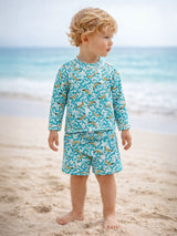 Child in a banana-patterned swimsuit stands on the beach, with ocean waves in the background.