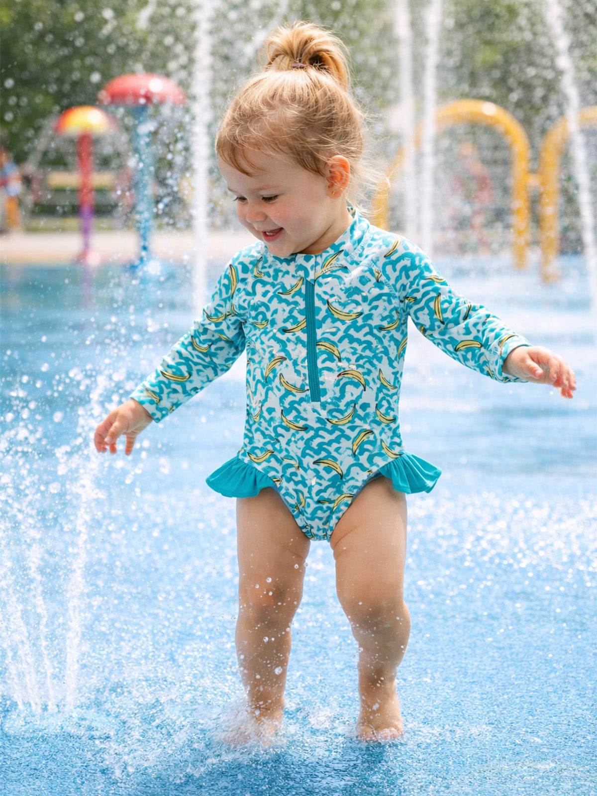 Toddler in a colorful swimsuit playing joyfully in a splash pad with water sprays around.