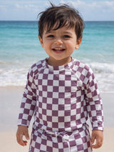 Smiling toddler in a checkered swimsuit on the beach, with ocean waves in the background.