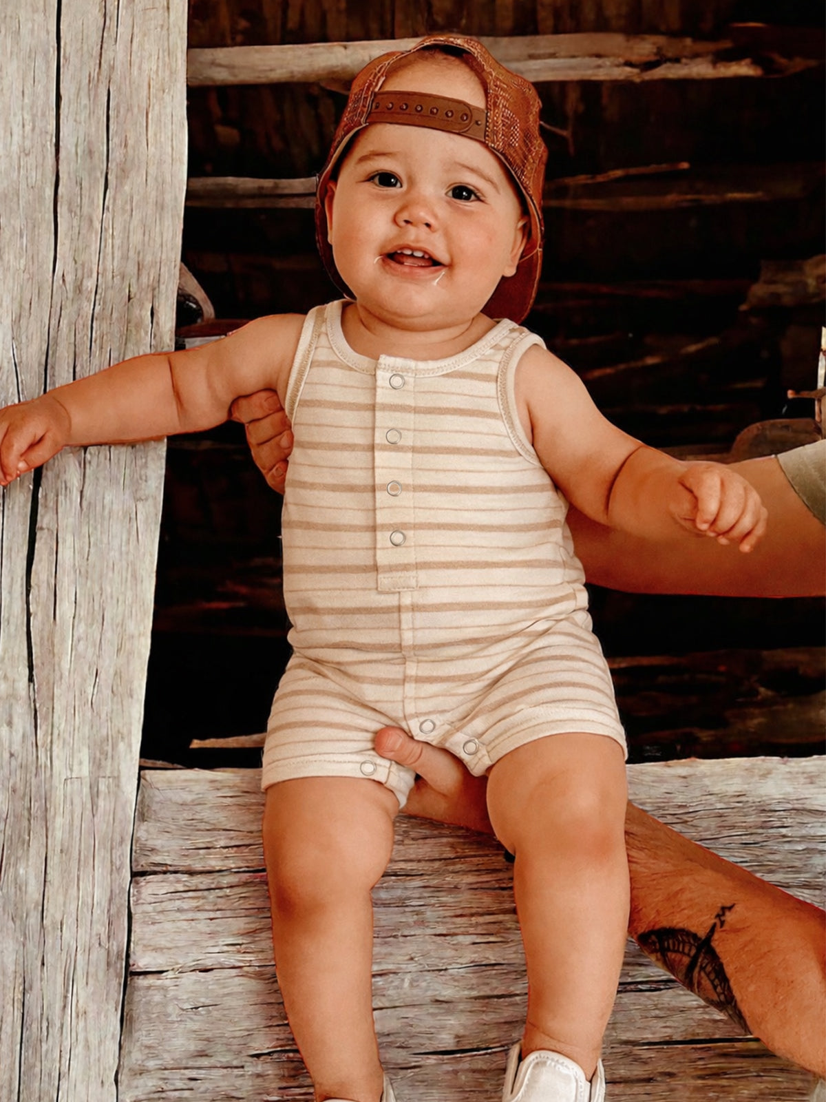 Smiling baby in a striped onesie and hat, sitting on a wooden beam, with a hand supporting them.
