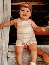 Smiling baby in a striped onesie and hat, sitting on a wooden beam, with a hand supporting them.