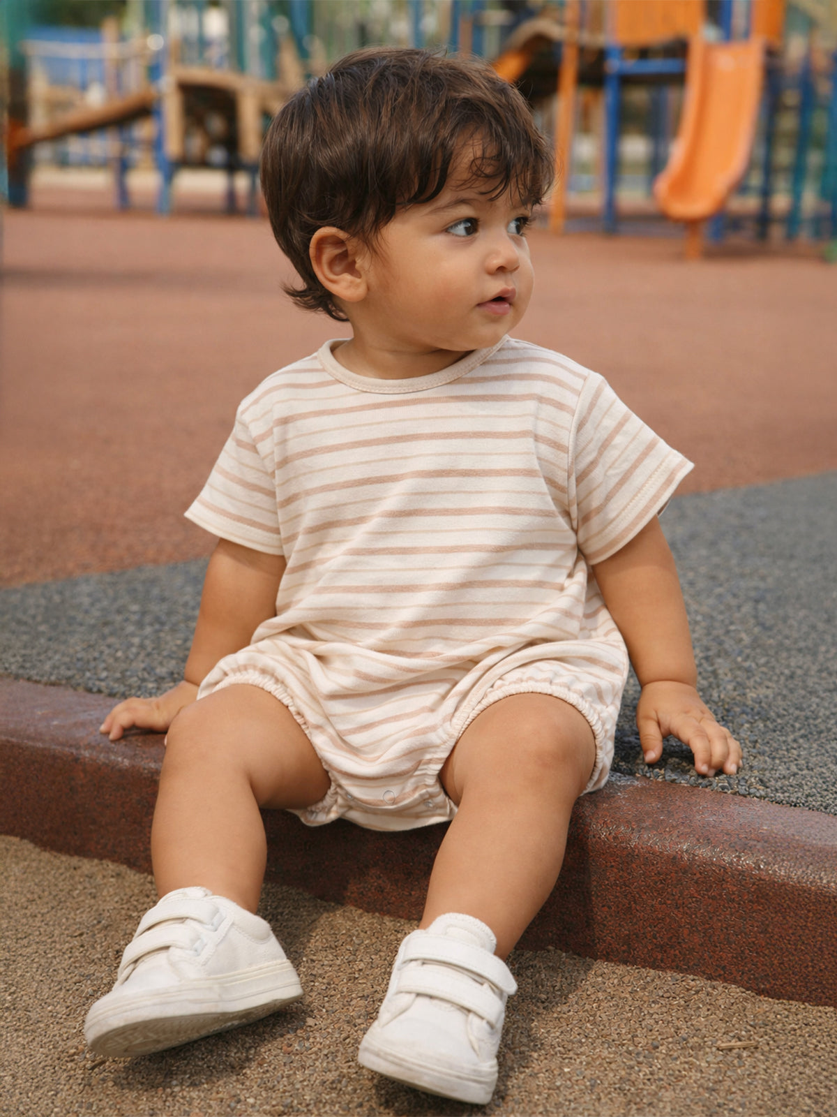 Child with curly hair sits on a playground surface, wearing a striped shirt and white shoes, looking to the side.