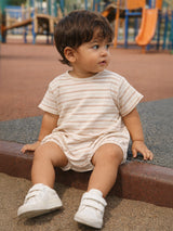Child with curly hair sits on a playground surface, wearing a striped shirt and white shoes, looking to the side.