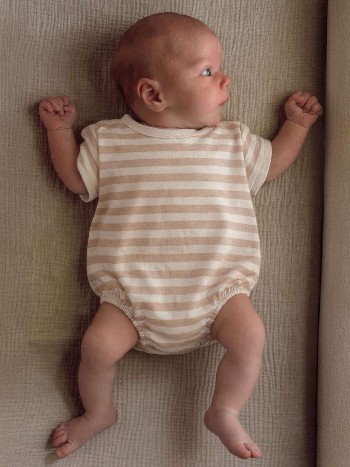 Infant in a striped onesie lying on a textured surface, looking to the side with a curious expression.