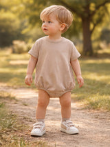 Toddler in a beige romper and sneakers stands on a path in a sunny, green park.