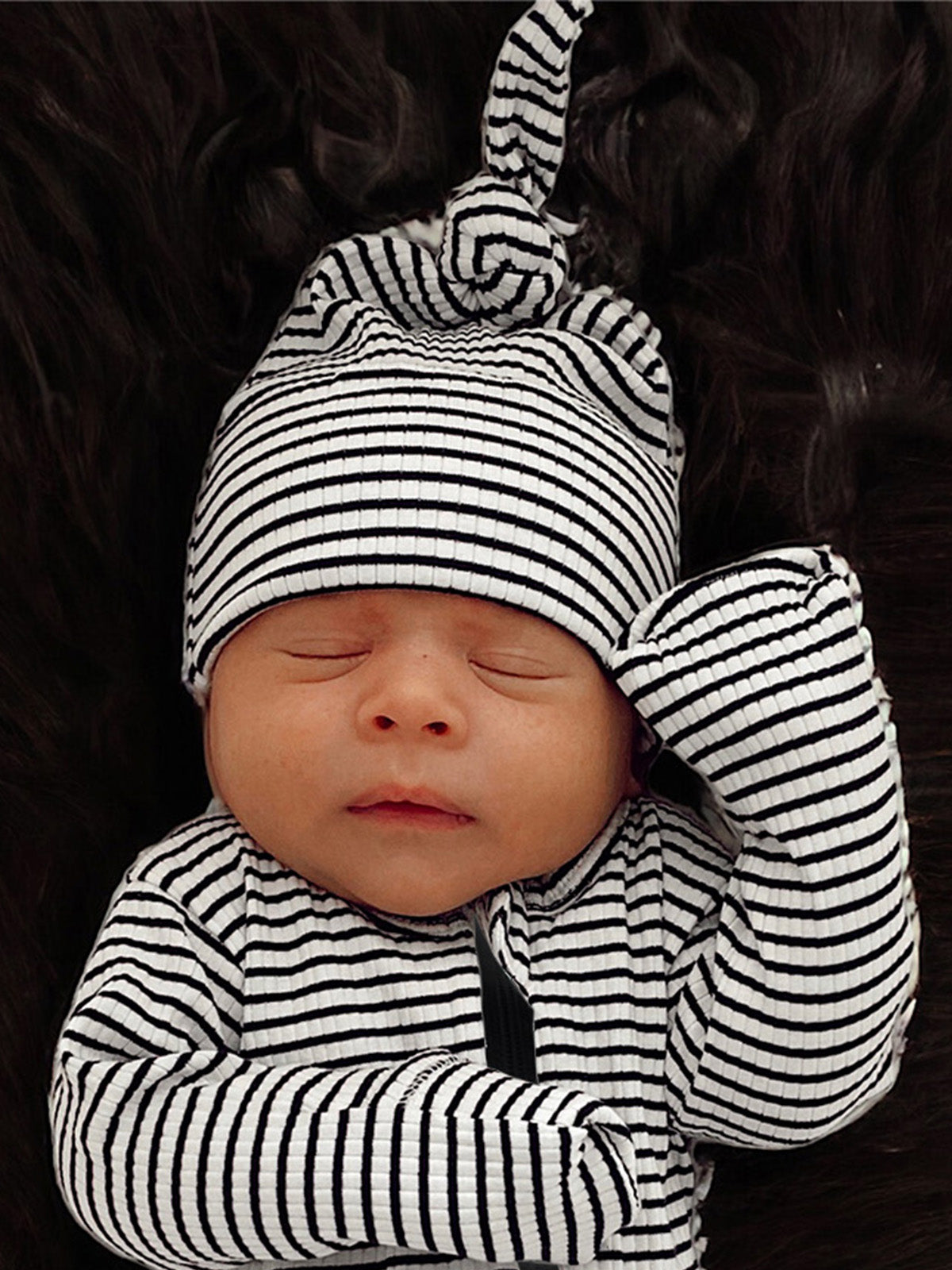 Newborn baby sleeping peacefully in black and white striped outfit and hat, against a dark textured background.