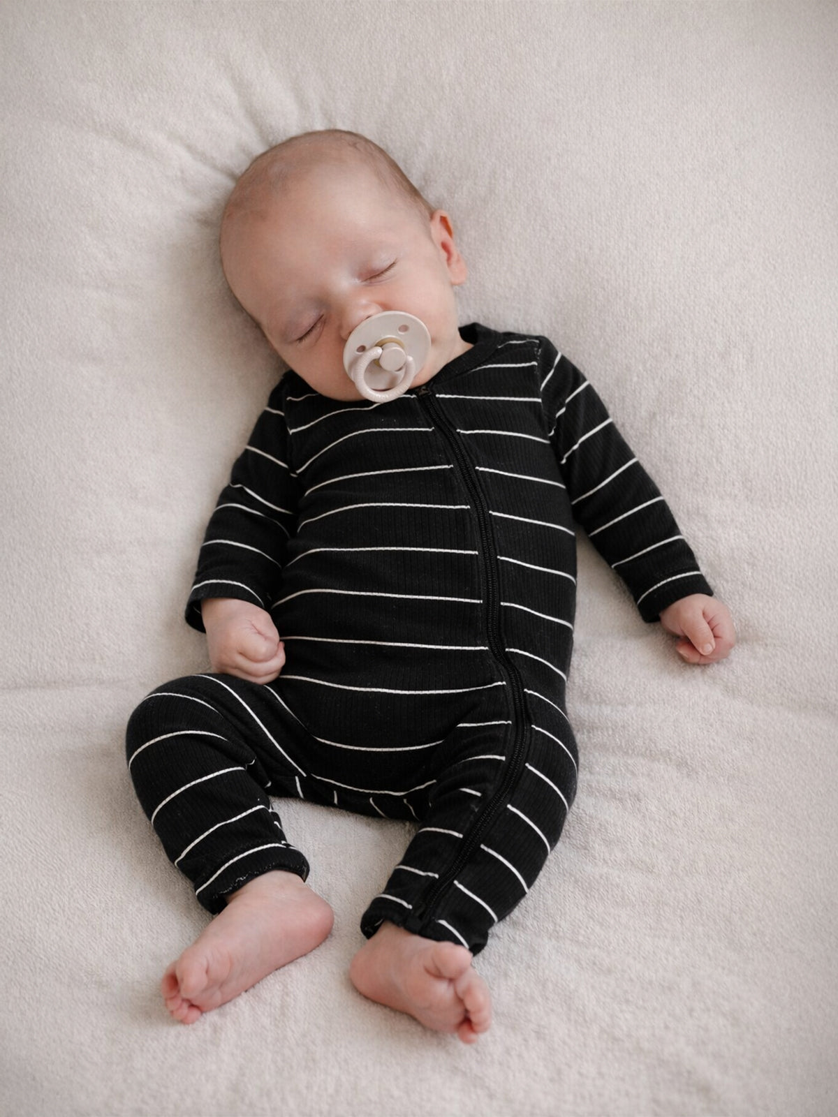 Sleeping baby in black and white striped pajamas with a pacifier, on a soft white blanket.
