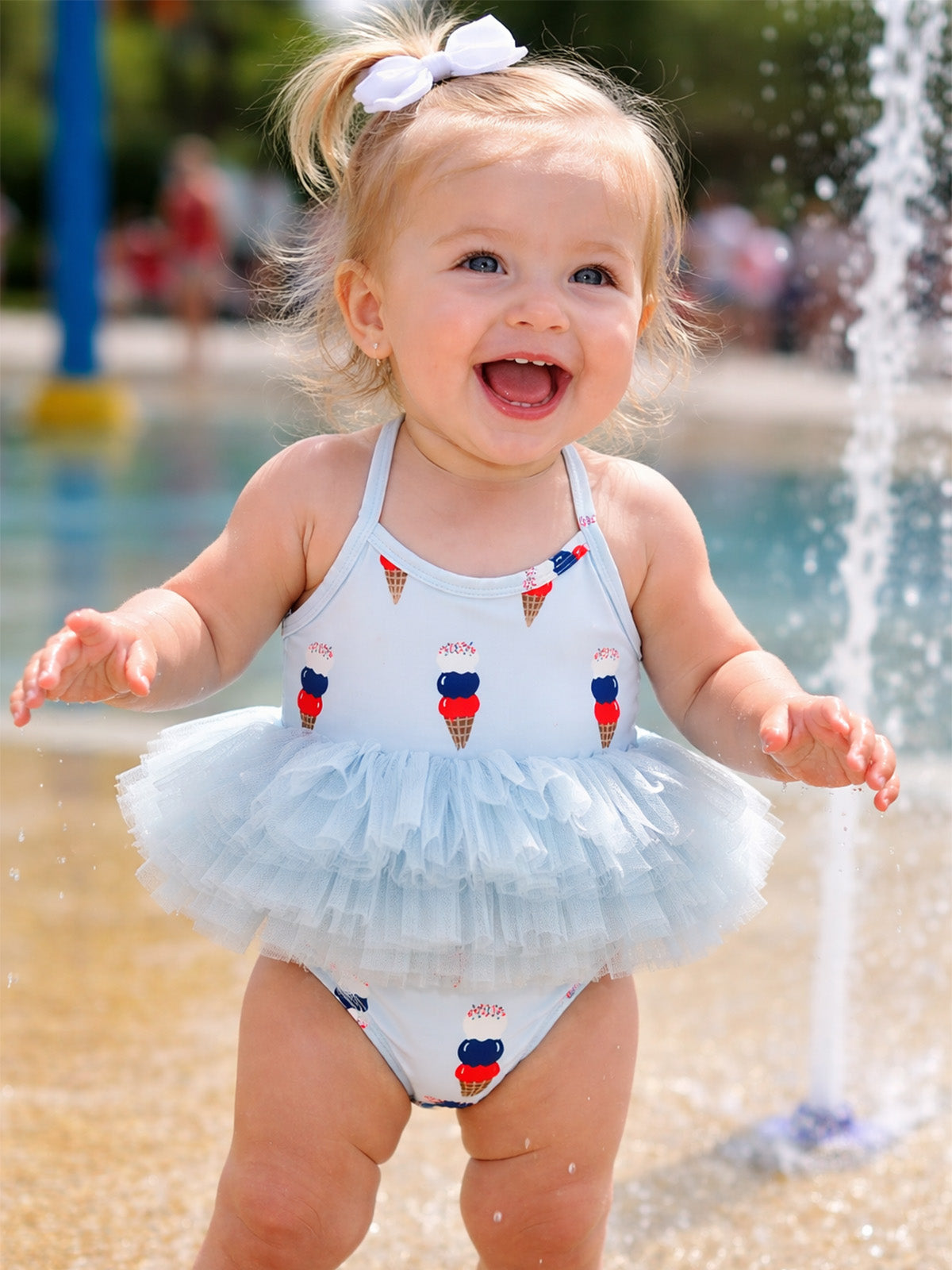 Smiling toddler in a playful ice cream print swimsuit, enjoying water splashes at a sunny outdoor location.