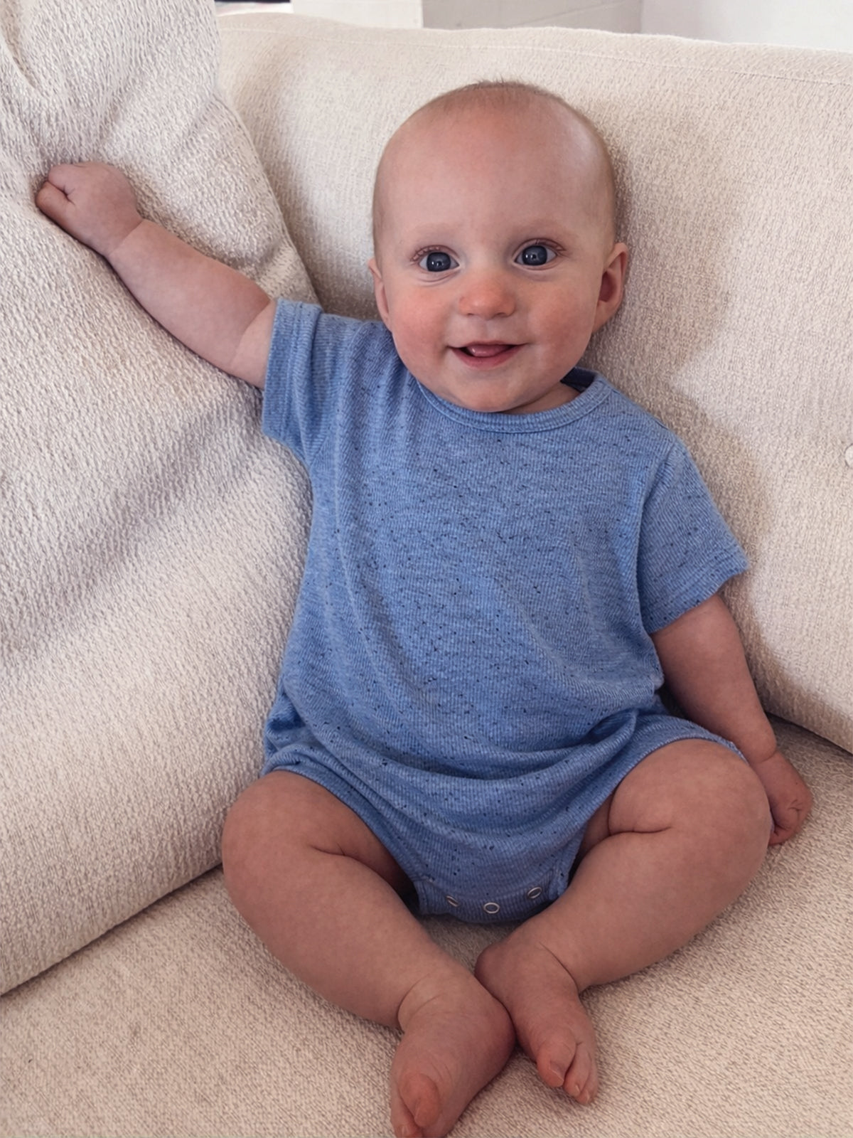 Smiling baby in a blue outfit sitting on a cream-colored couch, one arm raised playfully.