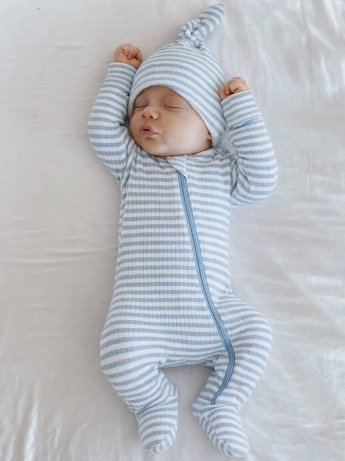 Infant sleeping peacefully in a blue and white striped onesie with a matching hat on a soft white surface.