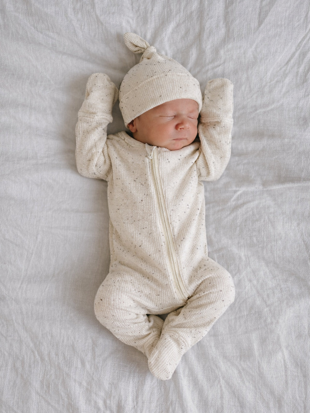 Newborn baby peacefully sleeping on a soft blanket, wearing a cream-colored outfit and a matching hat.