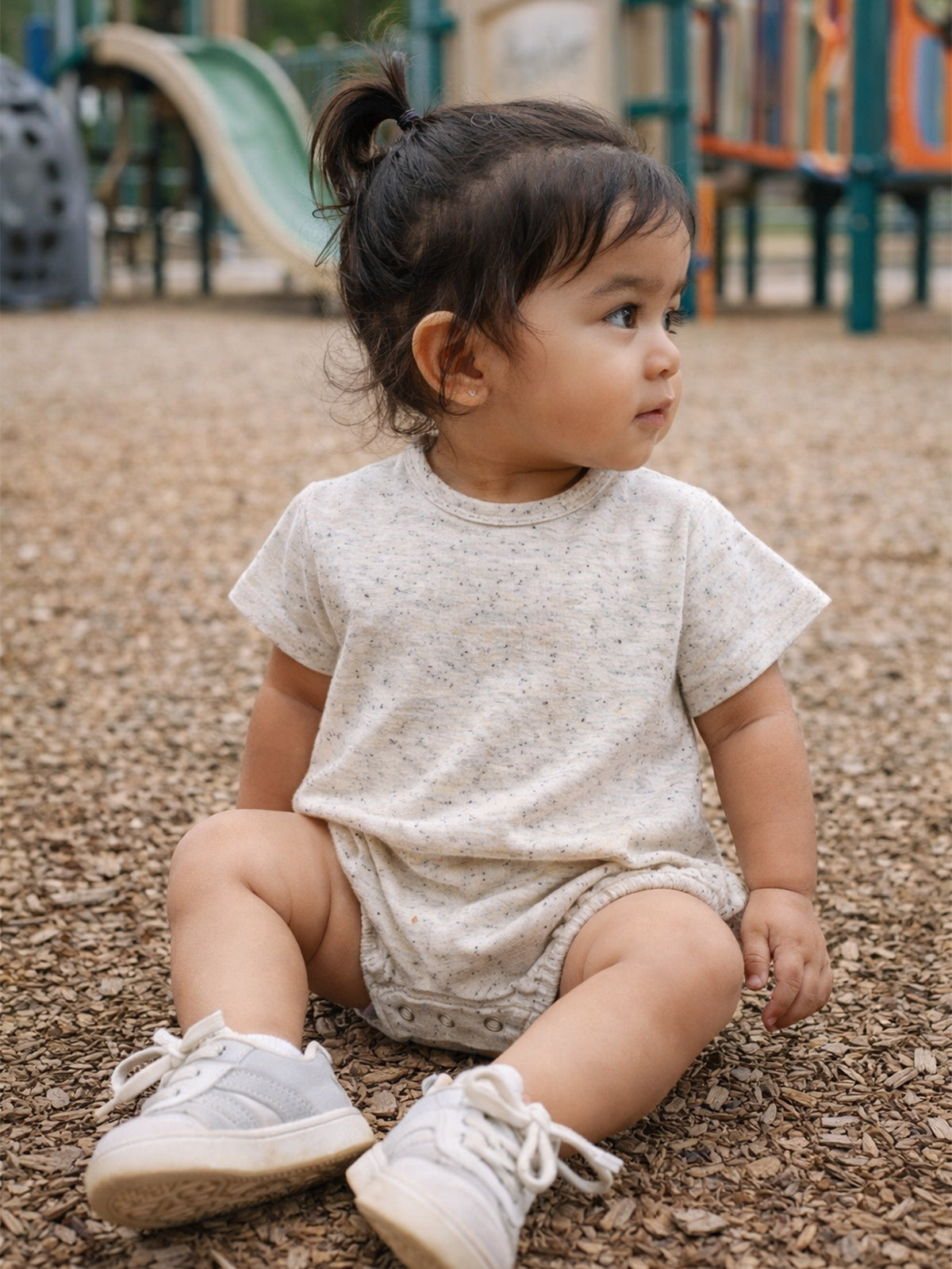 Toddler sitting on playground ground, wearing light shirt and shoes, looking to the side. Play equipment in background.