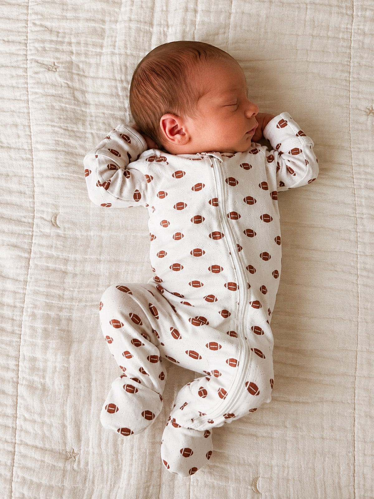 Newborn baby sleeping peacefully in a football-patterned onesie on a soft quilted blanket.