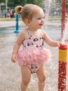 Toddler in a pink swimsuit with a ruffled skirt playing in a splash pad, smiling joyfully amidst water jets.