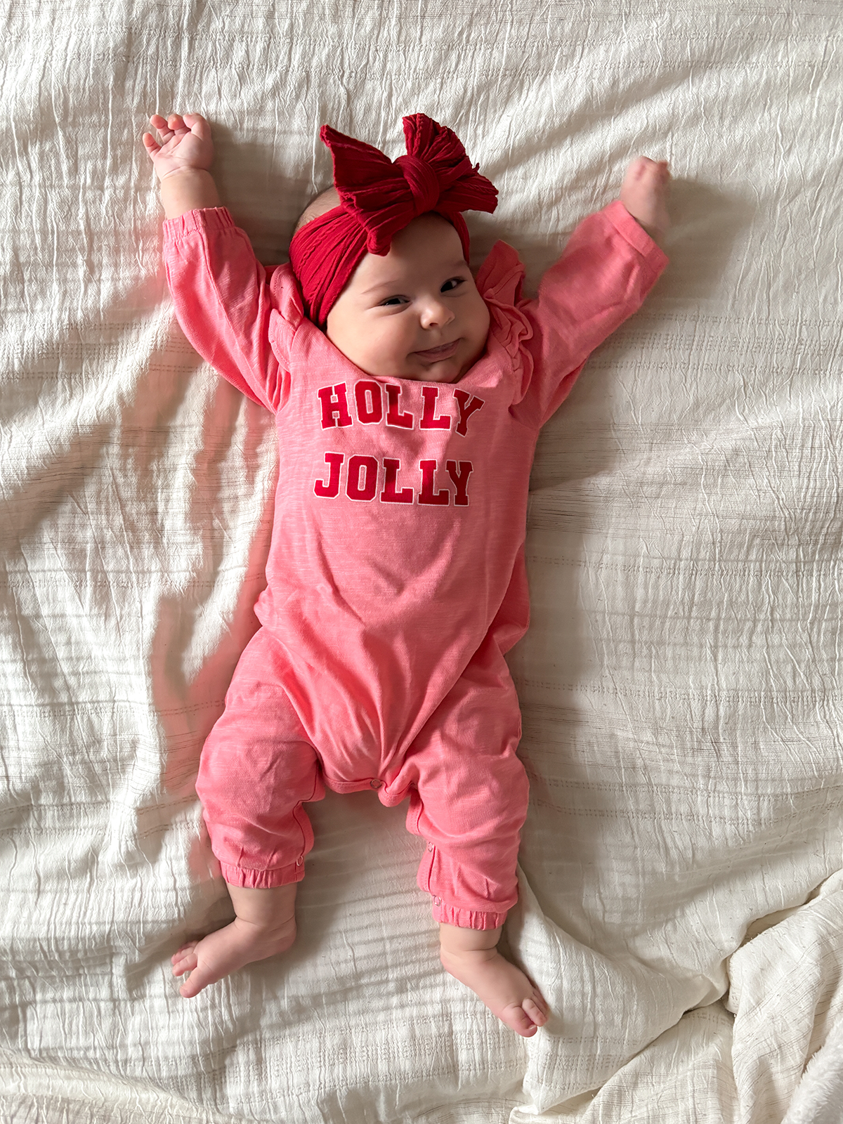 Baby lying on a white blanket, smiling, wearing a pink outfit with “HOLLY JOLLY” text and a red bow headband.