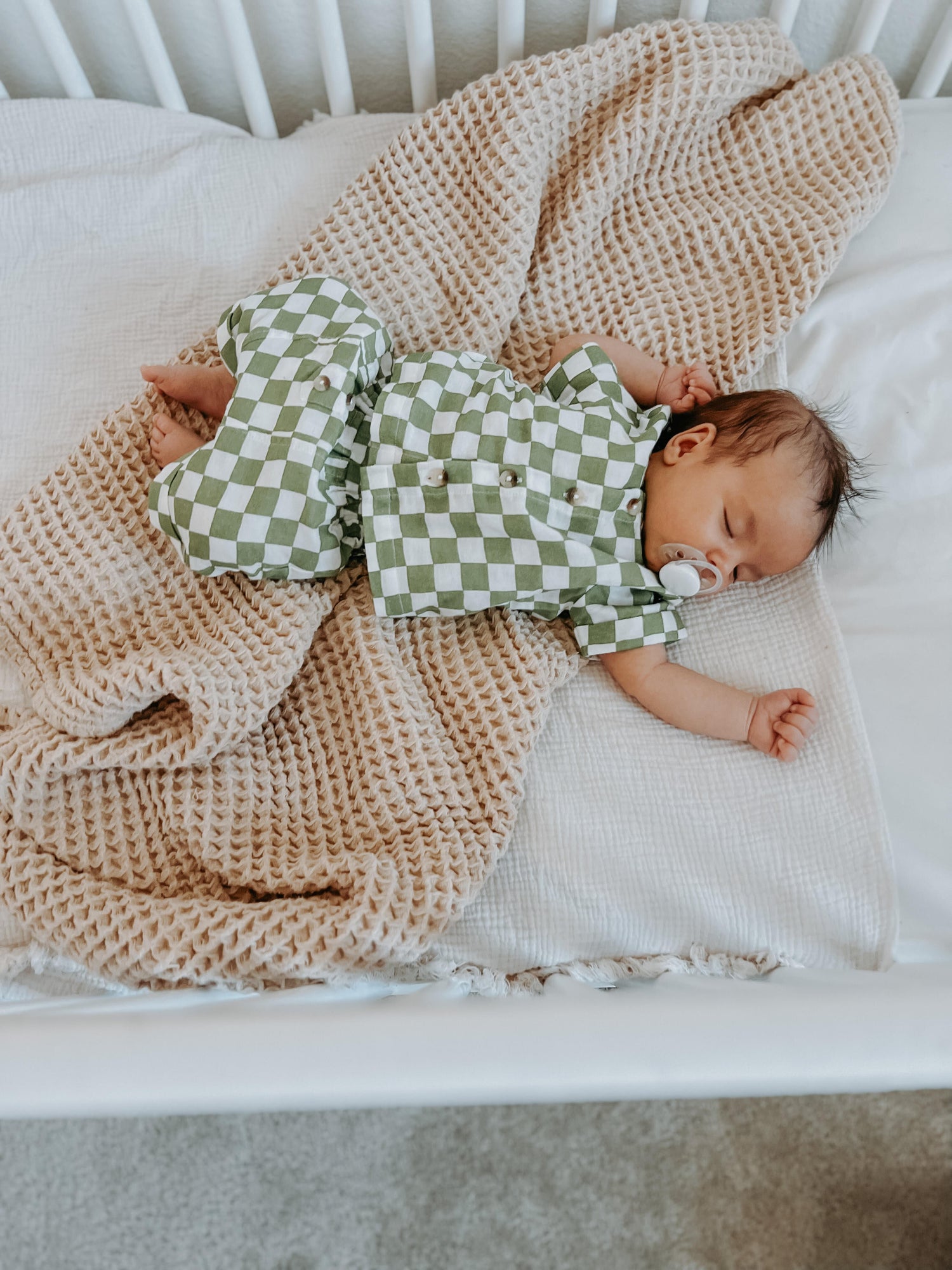 Baby peacefully sleeping in a crib, dressed in a green and white checkered outfit, surrounded by soft blankets.