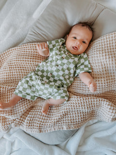 Baby in green and white checkered outfit lying on textured blanket, smiling at the camera.