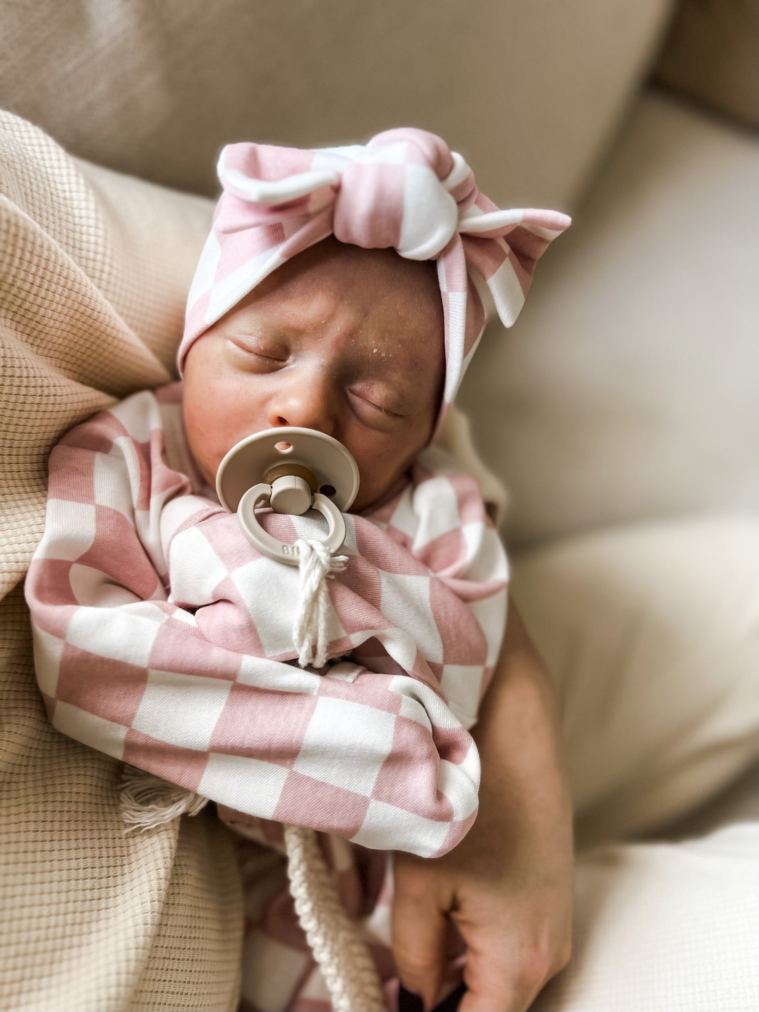 Newborn baby in pink checkered outfit and headband, peacefully sleeping with a pacifier in hand.