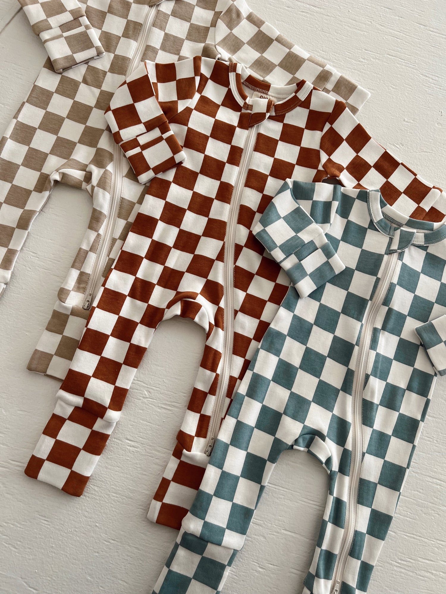 Three checkered baby onesies in brown, beige, and teal laid out on a white surface.