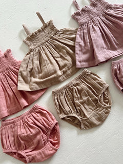 Assorted infants' linen tops and shorts in soft pink and beige hues on a light background.