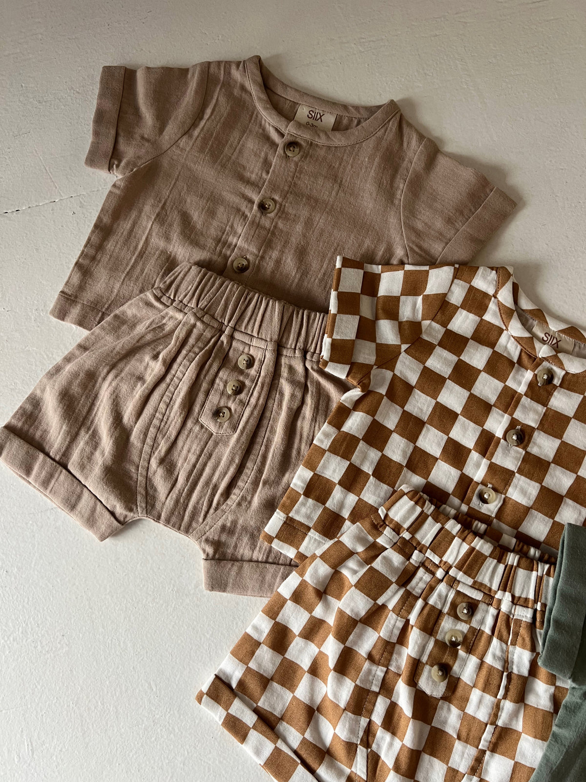 Two sets of children’s clothing: a beige short-sleeve top with shorts and a brown checkered set, all on a light surface.