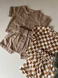 Two sets of children’s clothing: a beige short-sleeve top with shorts and a brown checkered set, all on a light surface.