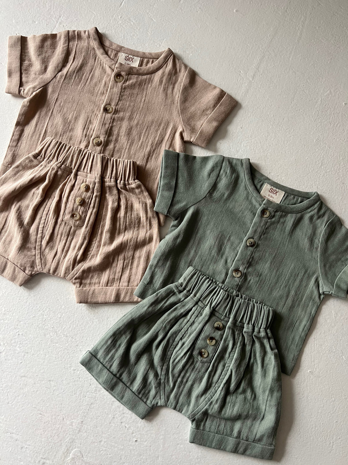 Two sets of infant romper outfits in beige and green with buttons, arranged on a neutral background.