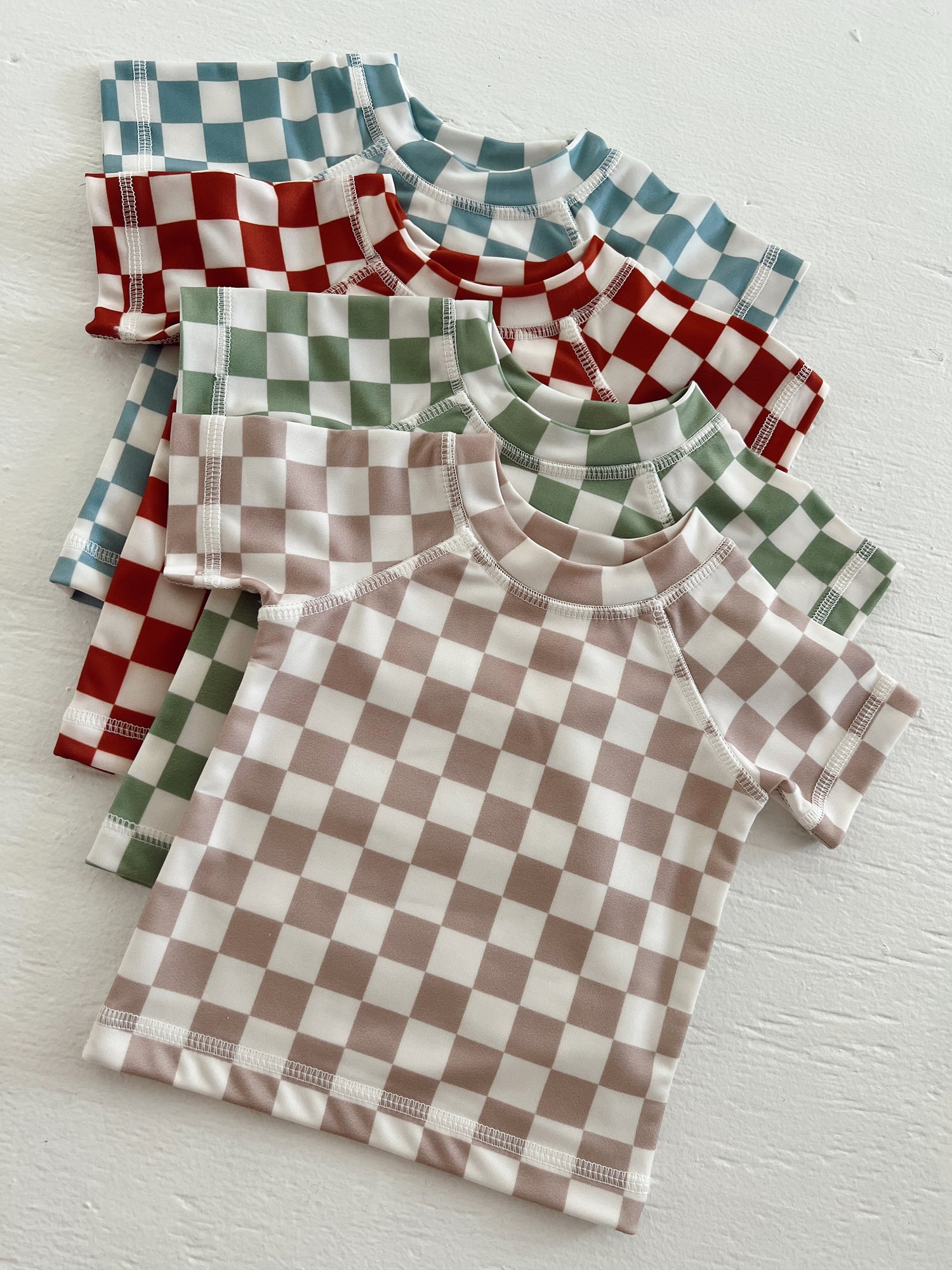Four short-sleeve checkered pattern shirts in blue, red, green, and beige, arranged on a light background.