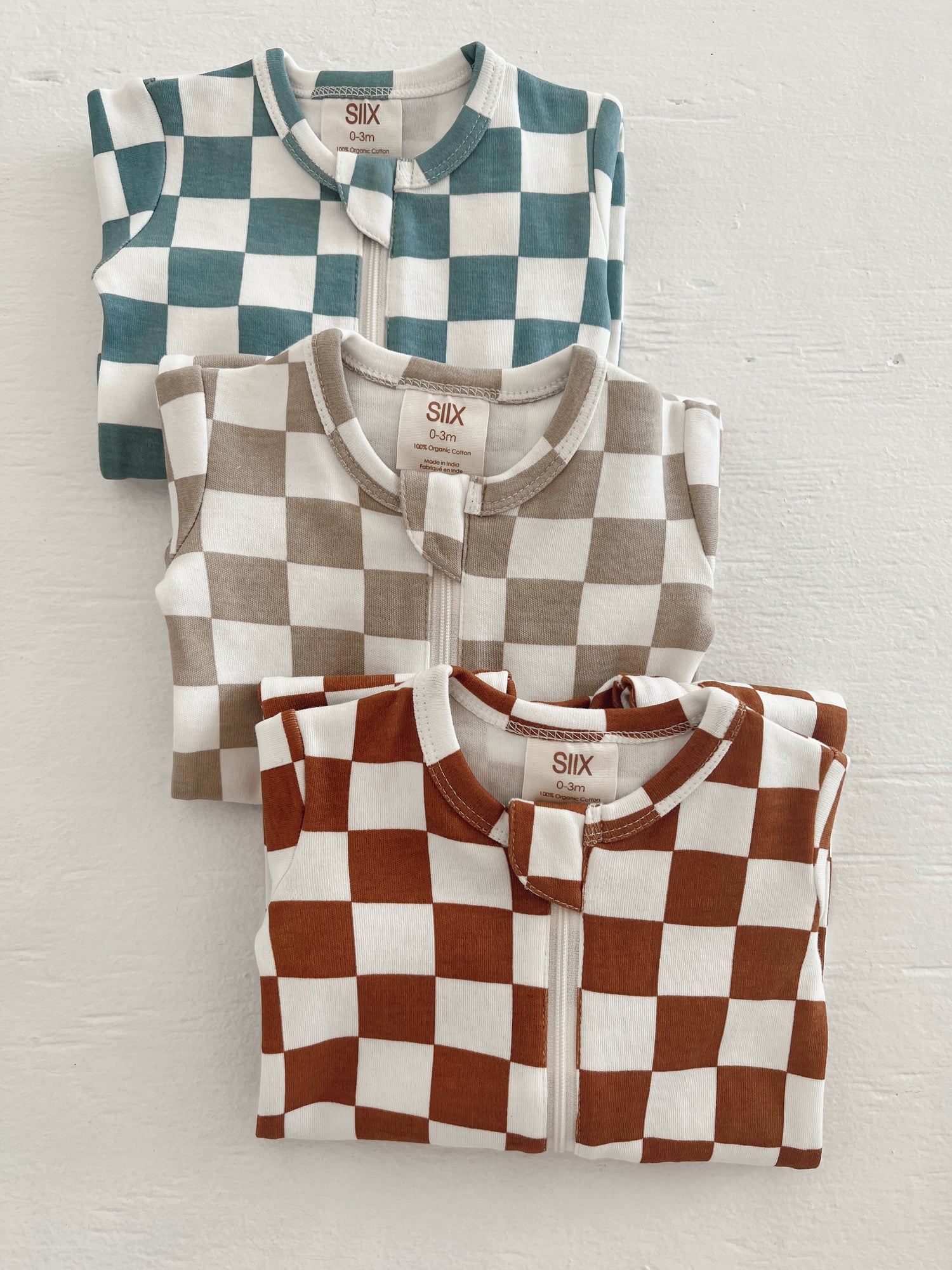 Three checkered infant onesies in teal, beige, and rust stacked against a white background.