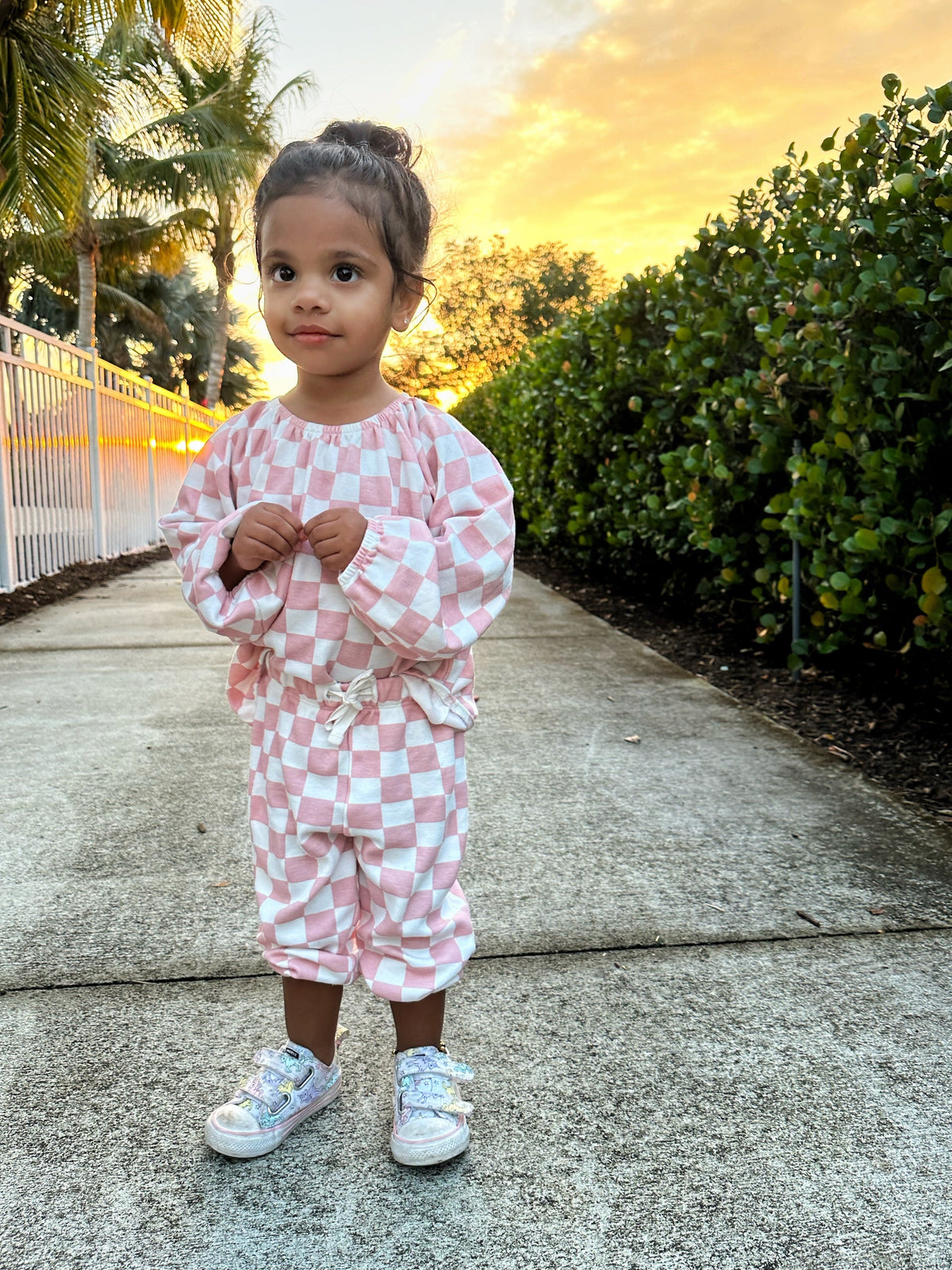 Child in pink checkered outfit stands on a sidewalk with a sunset backdrop and greenery.