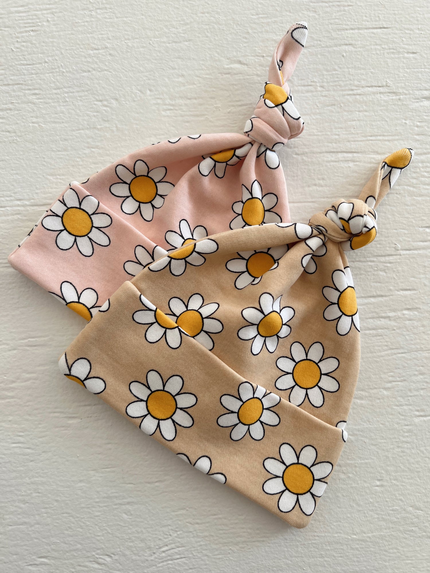 Two flower-patterned baby beanies, one in pink and one in beige, tied into knots.