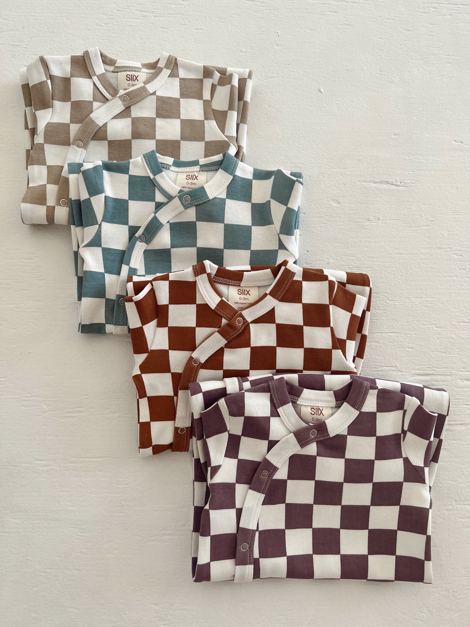 Four folded baby onesies in a checkered pattern: beige, teal, brown, and dark purple against a white background.