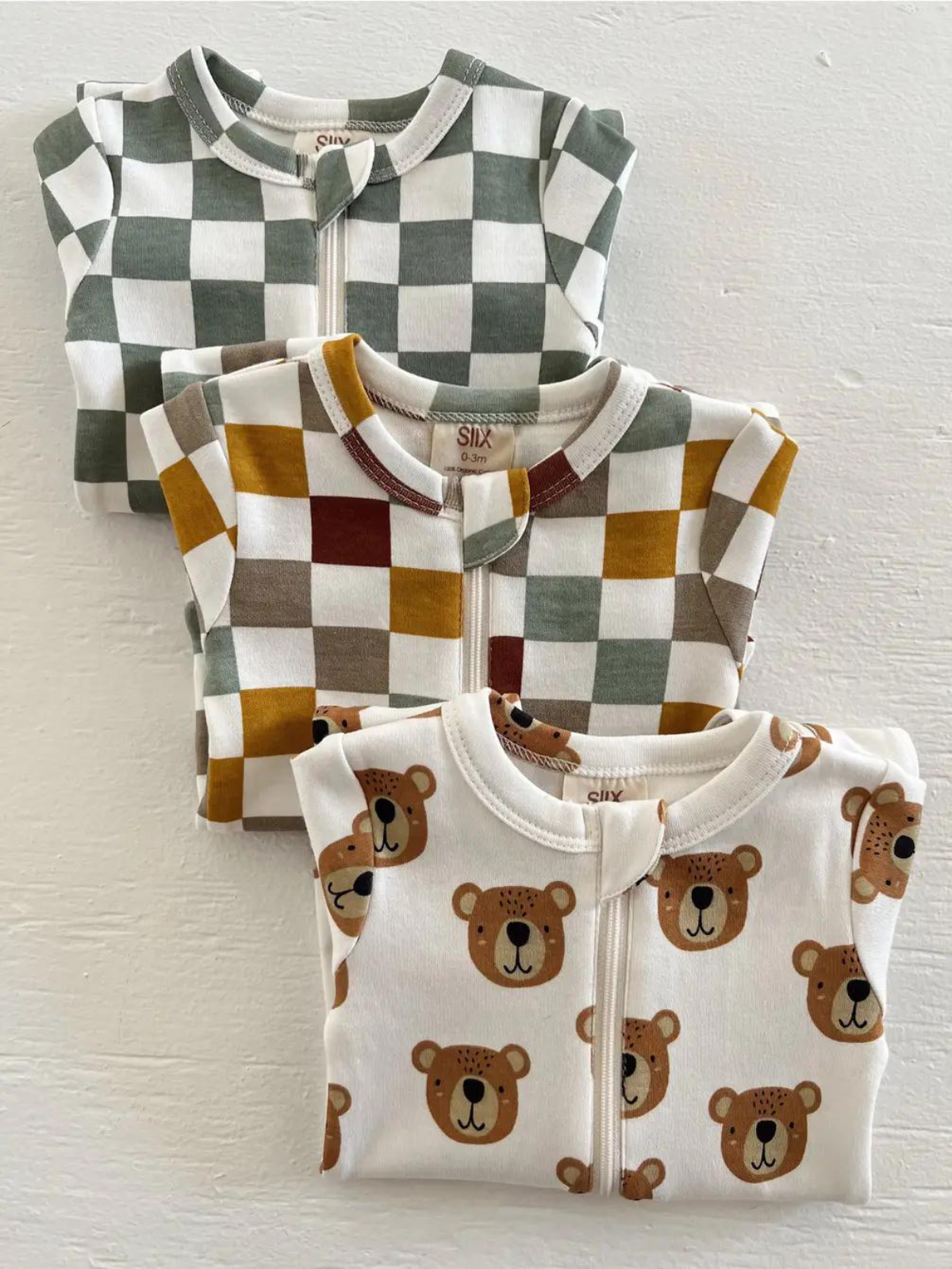 Three baby onesies featuring playful patterns: green and white checkers, mustard and brown checks, and bear faces.
