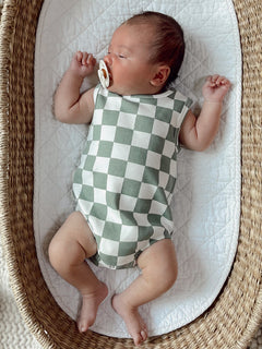 Baby wearing a green and white checkered onesie, lying in a woven basket with a pacifier.