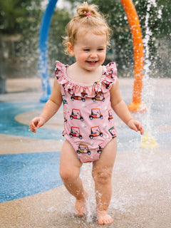 Smiling toddler in a pink swimsuit with ruffles, playing in a splash pad surrounded by colorful water features.