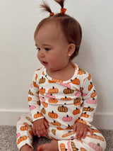 Toddler in pumpkin-patterned pajamas sitting on a carpet, with two playful pigtails against a white wall.