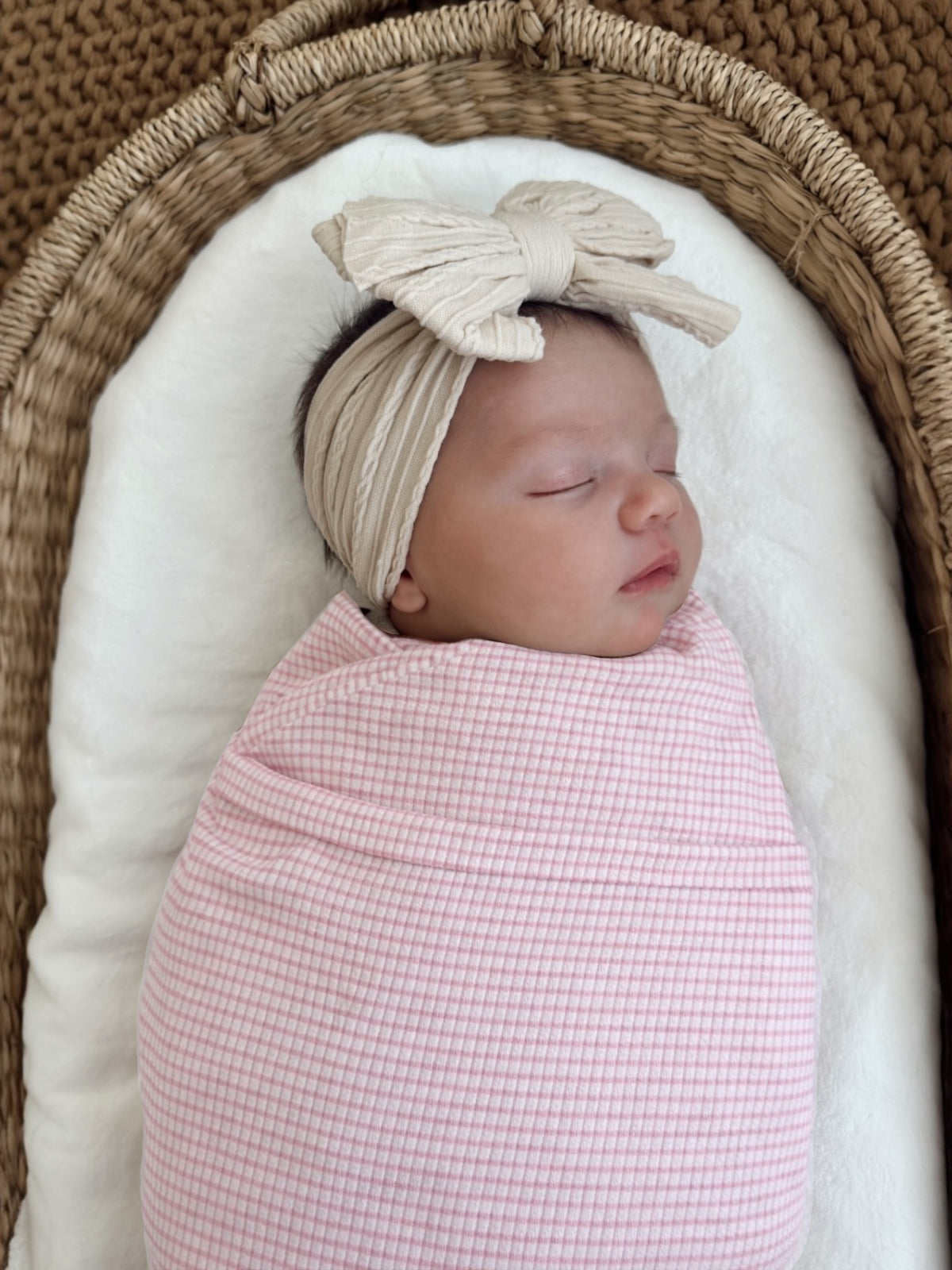 Sleeping baby wrapped in a pink checked blanket, wearing a beige bow headband, resting in a cozy basket.