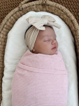 Sleeping baby wrapped in a pink checked blanket, wearing a beige bow headband, resting in a cozy basket.