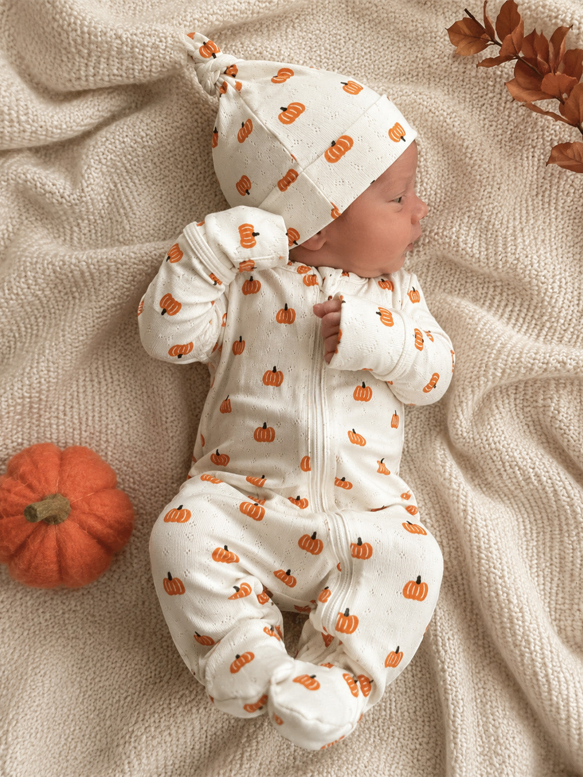 Newborn in pumpkin-printed outfit on a cozy blanket, next to a felt pumpkin and autumn leaves.