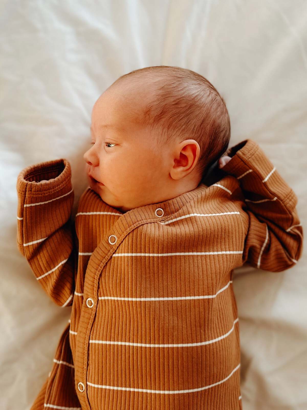 Newborn baby in a brown-striped outfit lying on a white blanket, gazing to the side.