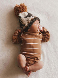 Sleeping baby in a brown striped onesie and a cozy knit hat with a pom-pom on a soft blanket.