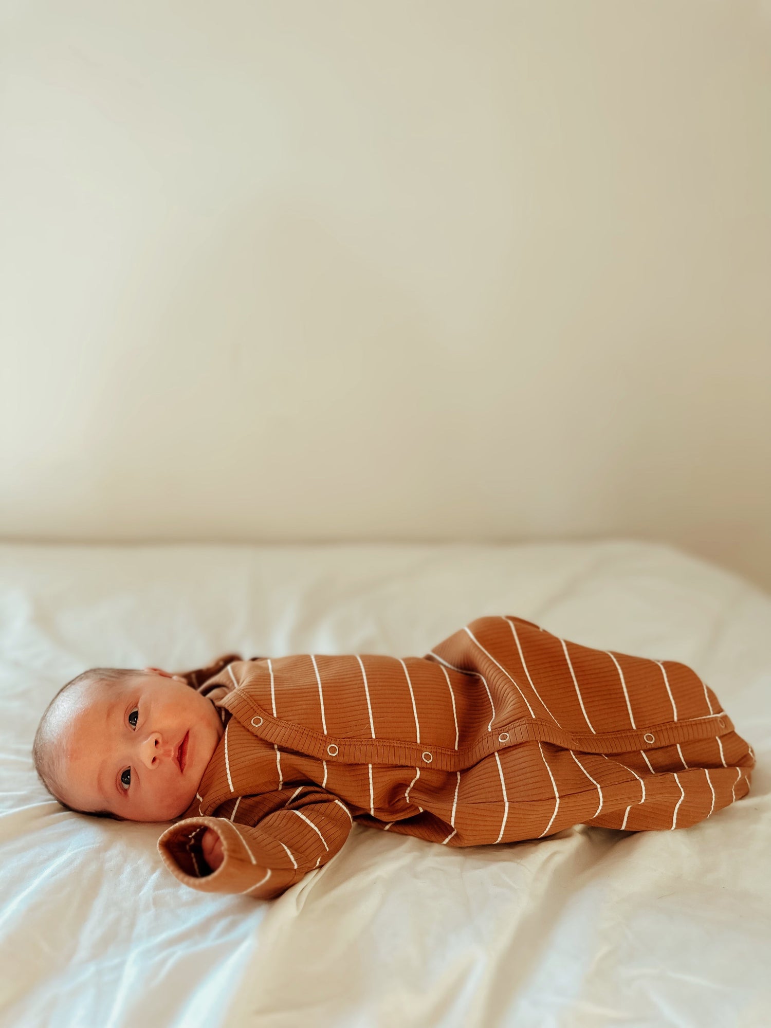 Baby in a brown striped outfit lying on a light-colored bedspread, looking curiously at the camera.