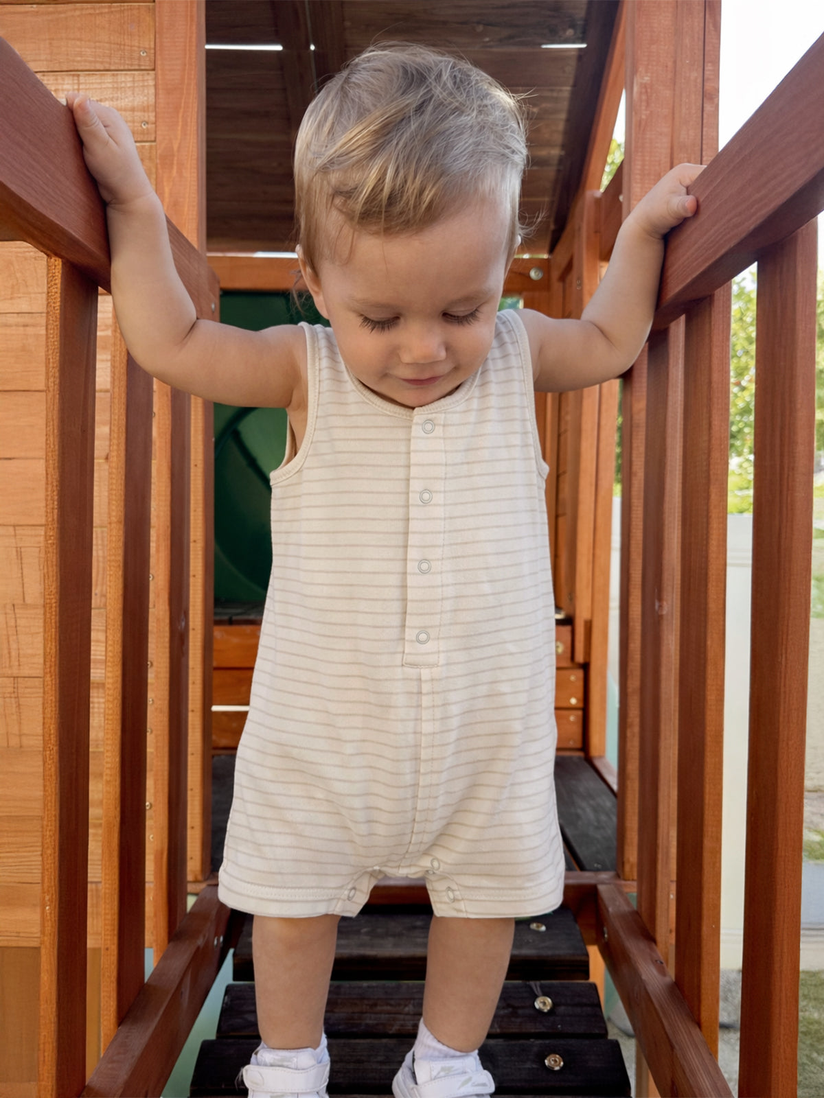 Toddler in a striped onesie standing on a wooden play structure, gripping the rail and looking down.