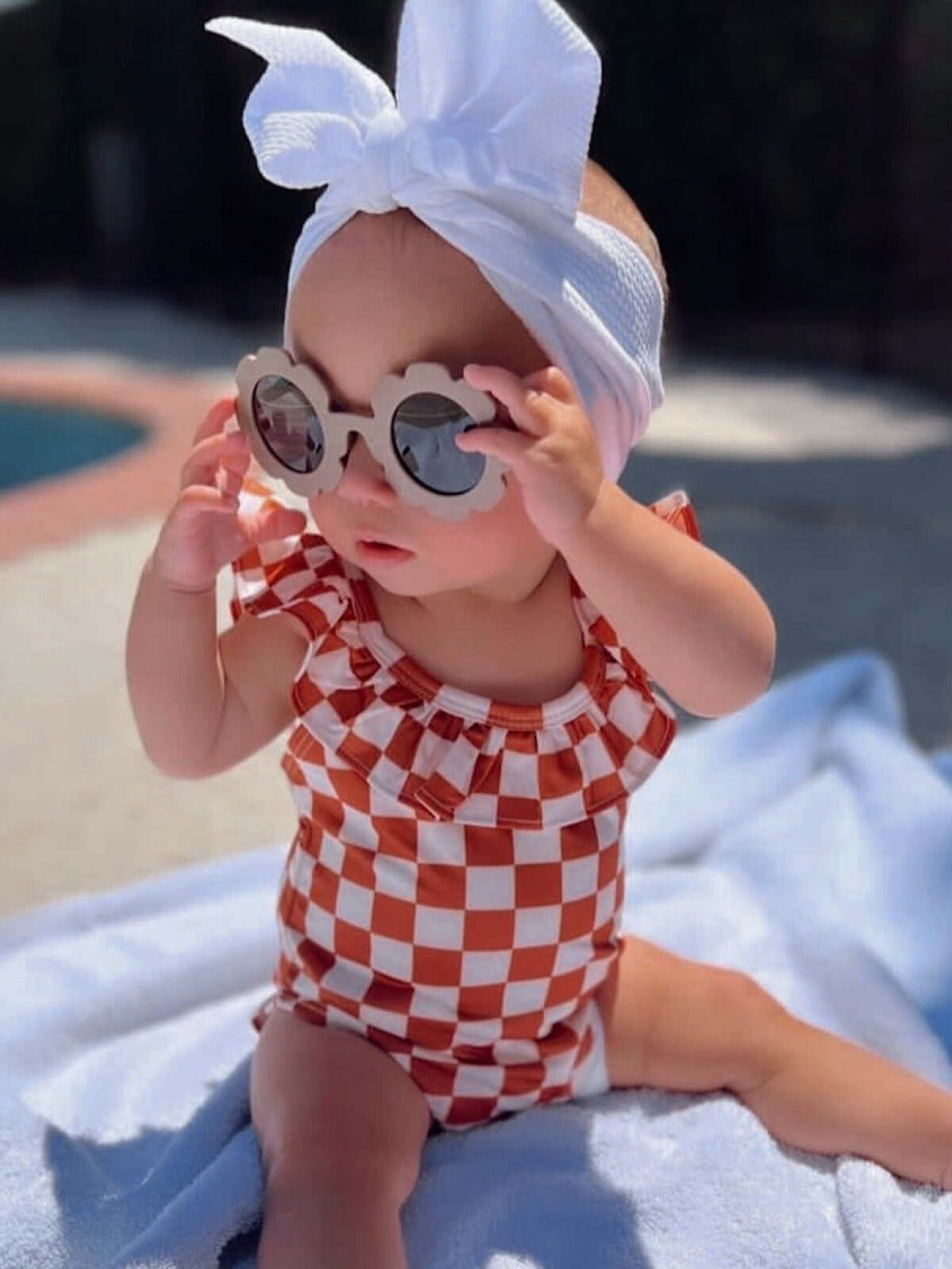 Baby in a checkered swimsuit and sunglasses sits on a towel by the pool, wearing a bow headband.
