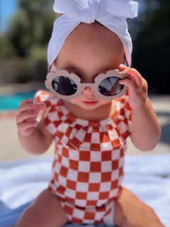 Baby wearing a checkered swimsuit and sunglasses, sitting on a blanket by the pool.