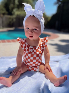 Baby in a checkered swimsuit and white bow sits on a towel by a poolside on a sunny day.