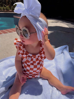Baby wearing a checkered swimsuit and sunglasses sits on a towel by the pool, with a large white bow in her hair.