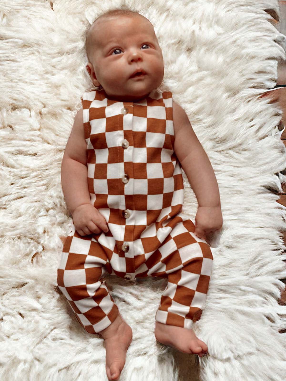 Baby in a brown and white checkered outfit sits on a fluffy white rug, gazing upward.