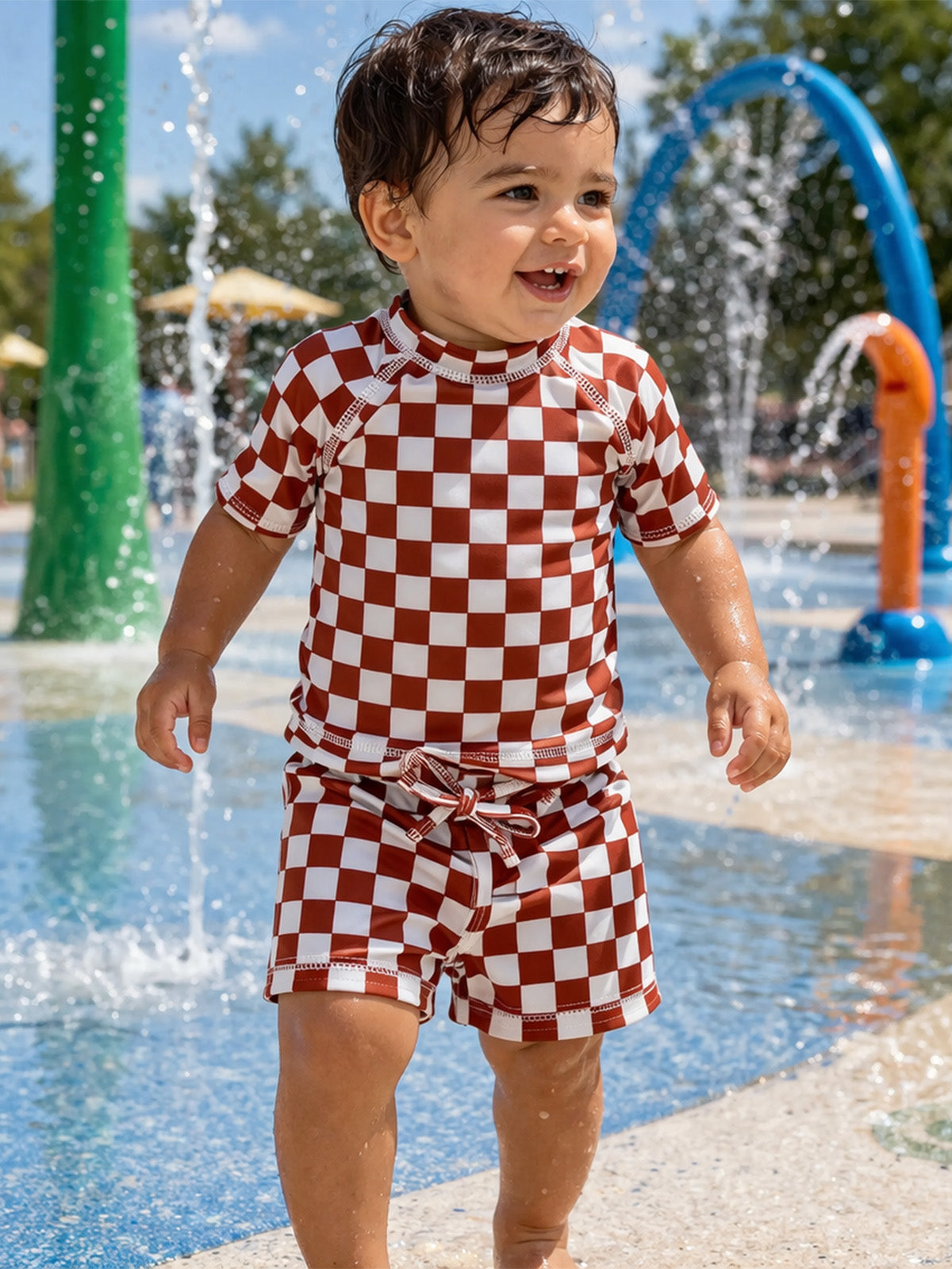 Child in red and white checkered swimwear happily playing in a splash pad with water fountains.
