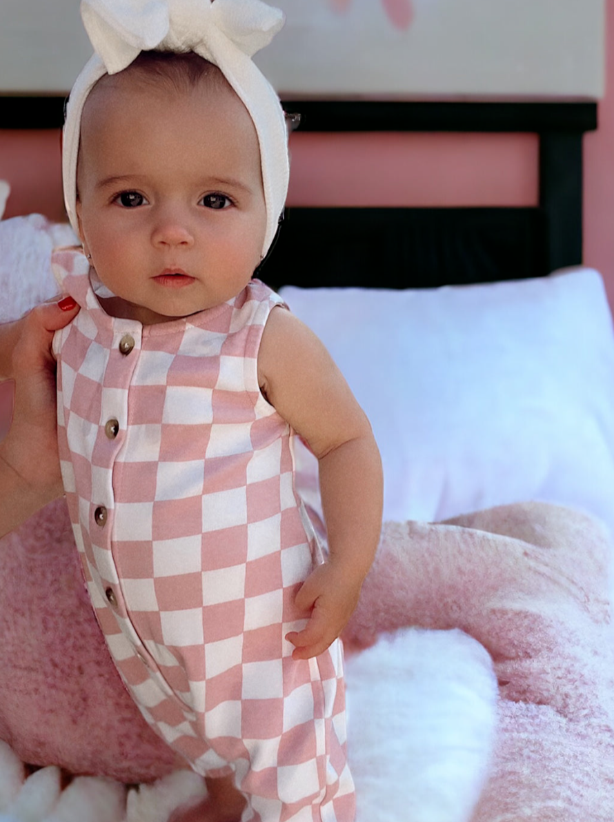 A baby wearing a pink and white checkered outfit and a white bow headband, sitting on a fluffy pink blanket.